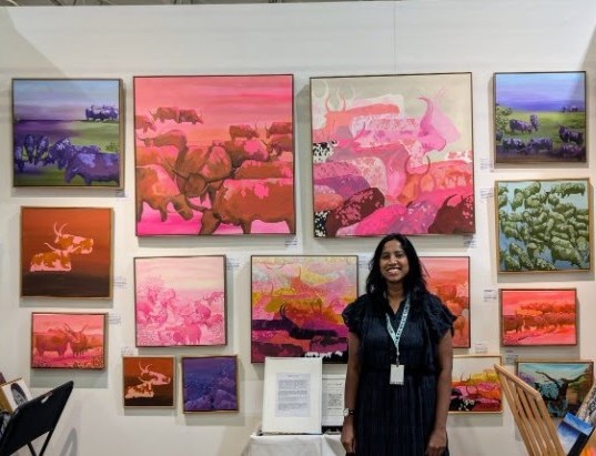 Bharathi standing in her exhibition booth surrounded by cattle paintings in pink and purple tones